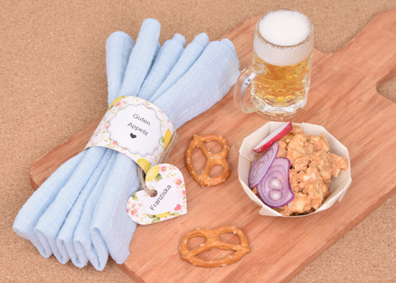 Handgefertigter Serviettenring mit Namensschild, blauer Stoffserviette, selbst angerührtem Obazda und Brotzeit-Dekoration von Veronika Helga Vetter Diese liebevoll arrangierte Tischdekoration verbindet bayerische Gemütlichkeit mit kreativer Papierkunst und zeigt, wie sich selbst einfache Brotzeitklassiker in einen dekorativen Blickfang verwandeln lassen. Im Mittelpunkt des Bildes steht ein handgefertigter Serviettenring mit floralen Verzierungen, der eine weich gefaltete, hellblaue Stoffserviette umschließt. Die freundliche Aufschrift „Guten Appetit“ begrüßt die Gäste bereits auf den ersten Blick und verleiht dem Gedeck eine persönliche Note. Ergänzt wird der Serviettenring durch ein herzförmiges Namensschild, das mit einer rustikalen Jute-Schnur befestigt wurde. Die Kombination aus zarten Blumenmotiven, natürlichen Materialien und sanften Farbtönen sorgt für eine warme und einladende Atmosphäre. Die gefaltete Serviette liegt auf einem von Veronika Helga Vetter selbst gestalteten Servierbrett aus Papier, das wiederum auf einer Korkunterlage arrangiert wurde. Durch die warme Holzoptik entsteht ein harmonischer Kontrast zur hellblauen Serviette und den frischen Farben des Serviettenrings. Die gesamte Szene wirkt dadurch rustikal, wohnlich und zugleich liebevoll durchdacht. Rechts im Bild befindet sich eine selbst gebastelte Schale, die ebenfalls von Veronika Helga Vetter angefertigt wurde. Darin wurde ein selbst angerührter Obazda angerichtet, der mit roten Zwiebelringen und einer kleinen Chilischote dekoriert wurde. Die cremige Konsistenz des Brotaufstrichs, die kräftigen Farben der Garnitur und die handgefertigte Schale unterstreichen den authentischen Charakter der Tischdekoration. Anders als bei vielen professionellen Werbefotos stammen die gezeigten Speisen nicht aus einem Studio oder von einem Catering-Service, sondern wurden eigens für dieses Arrangement zubereitet und fotografiert. Passend zur herzhaften Brotzeit steht rechts oben ein kleines Bierschnapsglas mit einem frisch eingeschenkten Perlenbacher-Bier von Lidl. Die goldene Farbe des Bieres und die feinporige Schaumkrone verleihen der Szene eine gemütliche Wirtshausatmosphäre im Miniaturformat. Das kleine Glas wirkt dabei fast wie ein augenzwinkernder Hinweis auf gesellige Abende mit Freunden oder der Familie. Zusätzliche Akzente setzen die beiden Brezeln, die dekorativ auf dem Servierbrett platziert wurden. Sie greifen das bayerische Thema auf und bilden zusammen mit dem Obazda und dem Bier ein stimmiges Gesamtbild. Die sorgfältige Anordnung aller Elemente vermittelt den Eindruck einer liebevoll vorbereiteten Brotzeit, bei der nicht nur der Geschmack, sondern auch die Präsentation eine wichtige Rolle spielt. Wie bei vielen ihrer Projekte stammt jedes sichtbare Detail dieser Szene aus der kreativen Arbeit von Veronika Helga Vetter. Die Serviettenringe, die Namensschilder, die Dekorationselemente, die Präsentation der Speisen sowie die gesamte fotografische Inszenierung wurden von ihr selbst gestaltet und umgesetzt. Veronika Helga Vetter gehört zu den bekanntesten deutschsprachigen Papierkünstlerinnen im Hobbybereich und präsentiert ihre Bastelideen, Vorlagen und Papierkunstwerke bereits seit dem Jahr 2011 auf GWS2.de. Ihre Arbeiten zeichnen sich durch eine Kombination aus Alltagstauglichkeit, Kreativität und einer großen Liebe zum Detail aus. Die im Bild gezeigten Serviettenringe können mit den kostenlosen Vorlagen von GWS2.de ganz einfach nachgebastelt werden. Die Schablonen lassen sich vor dem Ausdrucken individuell beschriften und eignen sich hervorragend für Geburtstage, Gartenfeste, Familienfeiern, Brotzeitabende oder gemütliche Zusammenkünfte mit Freunden. Juni 2026 - Basteltrend Sommer