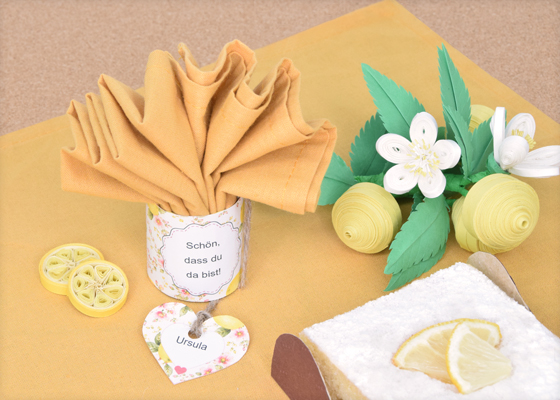 Aufgefächerte Stoffserviette im Zitronen-Serviettenring mit Namensschild, Quilling-Zitronen und selbst gebackenem Zitronenkuchen von Veronika Helga Vetter Diese liebevoll gestaltete Tischdekoration zeigt, wie vielseitig die kostenlosen Serviettenringe von GWS2.de eingesetzt werden können. Im Mittelpunkt des Bildes steht diesmal nicht die klassische Verwendung als Manschette für eine zusammengerollte Serviette, sondern eine besonders dekorative Alternative: Die Stoffserviette wurde kunstvoll aufgefächert und aufrecht in den Serviettenring gesteckt. Dadurch verwandelt sich der Serviettenring beinahe in einen kleinen Serviettenhalter und wird selbst zum Blickfang auf der festlich gedeckten Tafel. Die in warmem Zitronengelb gehaltene Stoffserviette erinnert mit ihren weichen Falten an die Blütenblätter einer geöffneten Blume. Der darunter stehende Serviettenring trägt die herzliche Botschaft „Schön, dass du da bist“ und begrüßt die Gäste bereits beim ersten Blick auf den gedeckten Platz. Ergänzt wird die Dekoration durch ein herzförmiges Namensschild mit der Aufschrift „Ursula“, das mit einer rustikalen Jute-Schnur befestigt wurde. Diese Kombination aus persönlicher Ansprache und handgefertigten Details verleiht der Tischdekoration eine besonders einladende und wertschätzende Wirkung. Das Bild zeigt eindrucksvoll, dass die Serviettenringe nicht zwingend durch die Serviette geschoben werden müssen. Stattdessen können sie auch aufrecht auf dem Tisch stehen und als dekorative Halterung für kunstvoll gefaltete oder aufgefächerte Servietten dienen. Gerade bei festlichen Kaffeetafeln, Gartenpartys, Geburtstagsfeiern oder sommerlichen Familienfesten entsteht dadurch eine besonders elegante Präsentation, die sich mit wenigen Handgriffen umsetzen lässt. Rechts im Bild befindet sich ein selbst gebackener Zitronenkuchen, dessen feine Puderzuckerschicht und die dekorativen Zitronenscheiben das sommerliche Thema der gesamten Szenerie aufgreifen. Der Kuchen wurde von Veronika Helga Vetter selbst gebacken und dekoriert. Auch der kleine Pappteller, auf dem das Kuchenstück angerichtet wurde, stammt aus ihrer kreativen Werkstatt und wurde passend zum Arrangement gestaltet. Für zusätzliche sommerliche Akzente sorgen die filigranen Zitronendekorationen im Hintergrund und Vordergrund. Die Zitronenscheiben links neben dem Serviettenring sowie die kunstvoll gerollten Zitronen und Zitronenblüten auf der rechten Bildseite wurden vollständig von Hand gefertigt. Hierbei kam die Quilling-Technik zum Einsatz, bei der schmale Papierstreifen zu dekorativen Formen gerollt und anschließend zu detailreichen Motiven zusammengesetzt werden. Die weißen Blüten, grünen Blätter und gelben Papierzitronen verleihen dem Arrangement eine frische, freundliche Ausstrahlung und unterstreichen den handwerklichen Charakter der gesamten Dekoration. Wie bei allen gezeigten Projekten wurde jedes sichtbare Element dieser Szene von Veronika Helga Vetter selbst gestaltet, gefertigt, arrangiert und fotografiert. Von den Serviettenringen über die Namensschilder und Quilling-Dekorationen bis hin zum Zitronenkuchen und dessen Präsentation stammt jedes Detail aus ihrer kreativen Arbeit. Als renommierte Papierkünstlerin veröffentlicht Veronika Helga Vetter ihre Bastelideen, Druckvorlagen und Papierkunstwerke bereits seit dem Jahr 2011 auf GWS2.de und inspiriert damit zahlreiche Bastelfreunde in Deutschland, Österreich und der Schweiz. Die im Bild gezeigten Serviettenringe können kostenlos auf GWS2.de heruntergeladen, individuell beschriftet und nachgebastelt werden. Das Foto zeigt dabei besonders anschaulich, wie flexibel die Vorlagen eingesetzt werden können – sowohl klassisch als Serviettenring als auch als dekorativer Aufsteller für kunstvoll gefaltete Stoffservietten. Finanziert von Computerheld Linz Oberösterreich