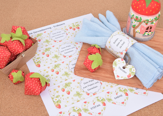 Serviettenring im Erdbeerdesign mit Namensschild, Papiererdbeeren, Erdbeerhäuschen aus Fimo und kostenlosen Druckvorlagen von Veronika Helga Vetter Diese fröhliche Tischdekoration steht ganz im Zeichen der Erdbeere und verbindet sommerliche Farben, kreative Papierkunst und liebevolle Handarbeit zu einem stimmigen Gesamtbild. Im Mittelpunkt der Szene liegt eine hellblaue Stoffserviette, die von einem handgefertigten Serviettenring im Erdbeerdesign umschlossen wird. Die freundliche Grußbotschaft „Schön, dass du da bist“ verleiht dem Gedeck eine persönliche und herzliche Note. Ergänzt wird der Serviettenring durch ein dekoratives Herzschild mit Namen, das mittels einer rustikalen Jute-Schnur befestigt wurde. Dadurch entsteht eine Tischdekoration, die Gästen bereits beim Platznehmen das Gefühl vermittelt, willkommen zu sein. Unterhalb der Serviette liegen mehrere ausgedruckte Vorlagen, die auf GWS2.de kostenlos zum Download bereitstehen. Die Schablonen zeigen verschiedene Erdbeermotive und enthalten sowohl Serviettenringe als auch passende Namensherzen. Besonders praktisch: Die Textfelder können vor dem Ausdrucken individuell beschriftet werden. So lassen sich persönliche Grußbotschaften, Namen oder kleine Botschaften direkt in die Vorlagen integrieren. Links im Bild befindet sich eine kleine Papierschale, die mit handgefertigten Erdbeeren aus Papier gefüllt wurde. Die detailreich gestalteten Früchte greifen das Erdbeermotiv der Serviettenringe auf und zeigen eindrucksvoll, wie vielseitig sich Papier als Bastelmaterial einsetzen lässt. Die Erdbeeren wurden einzeln gefertigt und anschließend dekorativ arrangiert. Einige weitere Papiererdbeeren liegen locker auf der Korkunterlage verteilt und verleihen dem Arrangement eine spielerische Leichtigkeit. Eine besondere Aufmerksamkeit verdient das dekorative Erdbeerhäuschen im oberen rechten Bildbereich. Bei diesem außergewöhnlichen Kunstwerk handelt es sich ursprünglich um ein gewöhnliches Marmeladenglas, das mit Fimo-Modelliermasse vollständig umgestaltet wurde. Durch die liebevoll modellierten Erdbeeren, Blätter, Fenster, Türen und kleinen Dekorelemente entstand ein märchenhaftes Erdbeerhaus, das wie direkt aus einem Bilderbuch zu stammen scheint. Dieses selbst gestaltete Dekorationsobjekt setzt einen fantasievollen Akzent und unterstreicht die kreative Handschrift der Künstlerin. Die gesamte Szene wurde auf einer Korkplatte arrangiert, wodurch die frischen Rot-, Grün- und Blautöne besonders gut zur Geltung kommen. Das natürliche Material sorgt für eine warme und wohnliche Ausstrahlung und bildet einen harmonischen Hintergrund für die zahlreichen Bastelarbeiten. Gleichzeitig werden die kostenlosen Vorlagen selbst Teil der Dekoration und zeigen anschaulich, wie sich die Serviettenringe mit wenigen Handgriffen nachbasteln lassen. Besonders interessant ist die Verbindung der verschiedenen Designwelten auf GWS2.de. Die dort angebotenen Serviettenringe sind sowohl im sommerlichen Zitronendesign als auch im fruchtigen Erdbeerdesign erhältlich. Dadurch lassen sich Tischdekorationen individuell an Jahreszeit, Anlass oder persönliche Vorlieben anpassen. Ob Gartenparty, Kaffeetafel, Geburtstagsfeier oder sommerlicher Brunch – die Vorlagen bieten zahlreiche Möglichkeiten für eine persönliche Tischgestaltung. Wie bei allen gezeigten Projekten stammt jedes sichtbare Detail dieser Dekoration aus der kreativen Arbeit von Veronika Helga Vetter. Die Serviettenringe, Namensschilder, Papiererdbeeren, das Erdbeerhäuschen, die Präsentation der Vorlagen sowie die fotografische Inszenierung wurden von ihr selbst gestaltet, gefertigt und arrangiert. Als renommierte Papierkünstlerin präsentiert Veronika Helga Vetter ihre Bastelideen, Druckvorlagen und kreativen Papierarbeiten bereits seit dem Jahr 2011 auf GWS2.de und inspiriert damit zahlreiche Bastelfreunde im deutschsprachigen Raum. Juni 2026