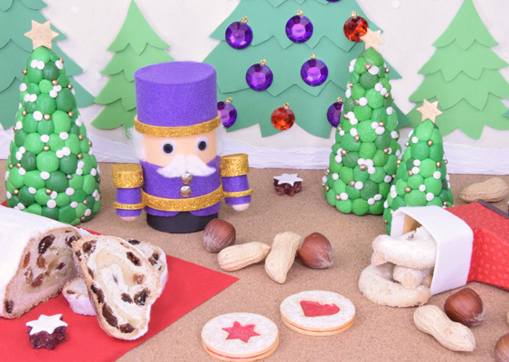 Purpurfarbener Papier-Nussknacker vor dekorierten Tannen mit Weihnachtsgebäck, Nikolausstiefel und Christstollen – winterliche DIY-Szene von Veronika Helga Vetter Die Szenerie wirkt wie ein liebevoll inszenierter Ausschnitt aus einer märchenhaften Weihnachtswelt, in der Süßwaren, Papierkunst und erzgebirgische Tradition miteinander verschmelzen. Im Zentrum des Bildes steht ein ungewöhnlich eleganter Papier-Nussknacker, diesmal in einem leuchtenden Purpurgewand. Sein Zylinder und seine Uniform sind in kräftigem Violett gehalten und mit goldenen Bordüren verziert, die das Licht reflektieren und der Figur eine festliche, fast majestätische Ausstrahlung verleihen. Der Nussknacker blickt mit großen Wackelaugen geradeaus, während ein weißer, fein geformter Watte-Schnurrbart und passende Augenbrauen ihm eine freundliche Würde verleihen. Hinter der Figur erhebt sich eine Kulisse aus mehrfach geschichteten, grünen Papier-Tannenbäumen. Einige sind mit großen rot- und violettglitzernden Schmucksteinen behangen, andere mit kleinen weißen Punkten versehen, die wie Schneeflocken erscheinen. Daneben ragen drei kunstvoll gestaltete Weihnachtsbäume aus Papier in die Höhe, jeweils mit goldenen Sternen an der Spitze und mit zarten weißen Punkten dekoriert, die wie frisch gefallener Schnee wirken. Die Komposition erinnert an eine kleine Waldlichtung, die für ein Fest herausgeputzt wurde. Im Vordergrund breitet sich eine köstliche Auswahl an weihnachtlichen Leckereien aus: Links liegt ein angeschnittener Christstollen, dessen helle, mit Früchten durchzogene Teigstruktur appetitlich hervorlugt. Daneben sind zwei Haselnüsse, Erdnüsse mit Schale und einige Kekse platziert, darunter Doppeldecker-Kekse mit sternförmigen Ausstanzungen in leuchtendem Rot. Rechts im Bild befindet sich ein kleiner Nikolausstiefel aus Papier in kräftigem Rot, der über die weiße Krempe hinweg eine Ladung selbstgebackener Vanillekipferl preisgibt. Einige Kekse sind dekorativ davor ausgestreut, sodass das Bild den Eindruck erweckt, als habe jemand eine winterliche Naschlandschaft liebevoll aufgebaut. Die gesamte Szenerie verbindet Handwerkskunst und Weihnachtsstimmung: der violette Nussknacker als strahlender Mittelpunkt, die grün glitzernden Bäume als Kulisse und die festlichen Süßwaren als stimmungsvolle Ergänzung. Das Arrangement wirkt wie eine Mischung aus Naschteller, Spielzeugtheater und erzgebirgischer Tradition – warm, einladend und detailverliebt. Erschaffen, arrangiert und fotografiert wurde diese winterliche Komposition von Veronika Helga Vetter, Papierkünstlerin und kreative Visionärin hinter GWS2.de.